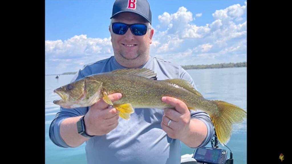 A smiling man, possibly Mike Frisch, wearing sunglasses and a baseball cap holds a large fish while standing on a boat, with calm water and a partly cloudy sky in the background.