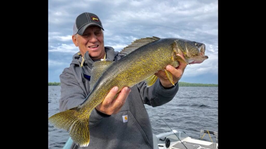 A smiling man, mike frisch, in a gray hoodie and cap holds a large yellow walleye fish with both hands while standing on a boat on a lake under a cloudy sky.