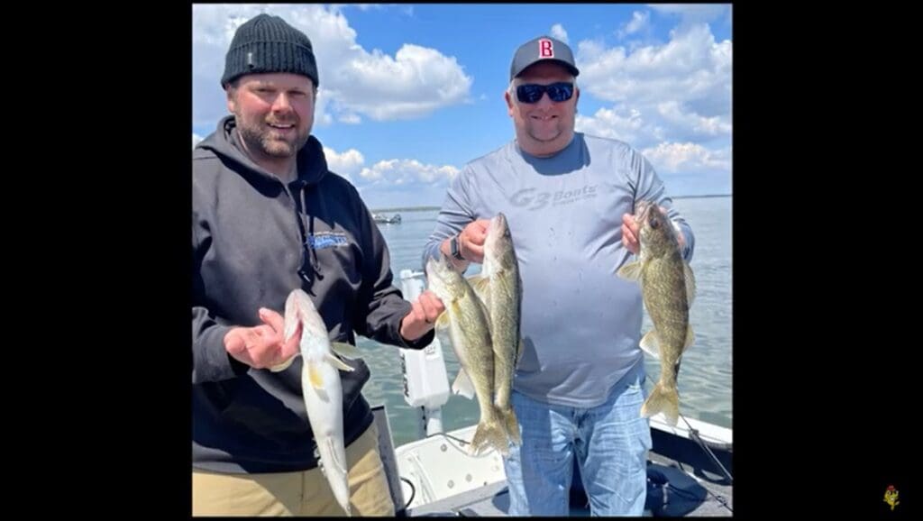 Two men stand on a boat, proudly holding up several large fish they caught while fishing, smiling at the camera. The sky is partly cloudy, and calm water stretches out behind them. Both are dressed in casual outdoor clothing.