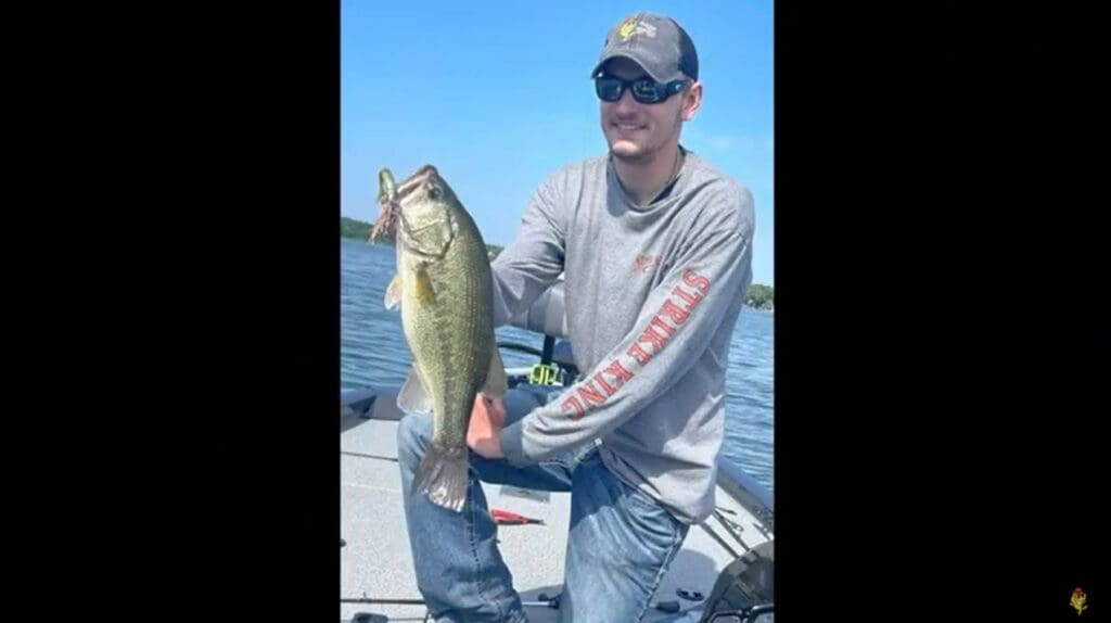 A man in sunglasses and a blue cap smiles while proudly holding his catch—a large fish—on a boat, with water and sky in the background.