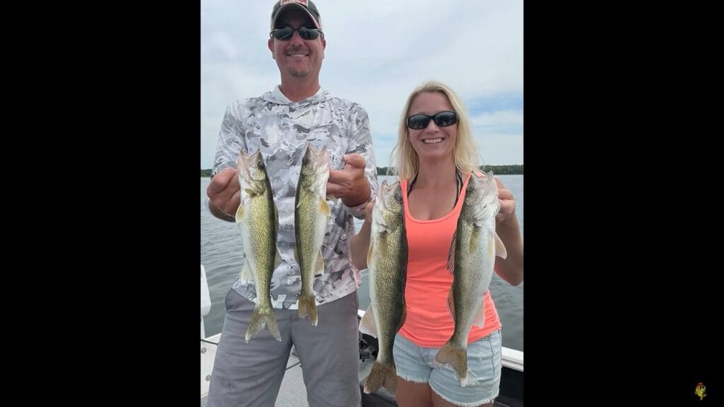 A man and a woman on a boat each hold up two walleye, smiling at the camera. The man wears a camo shirt, cap, and sunglasses; the woman sports sunglasses, a neon tank top, and shorts. Water and sky stretch into the background.