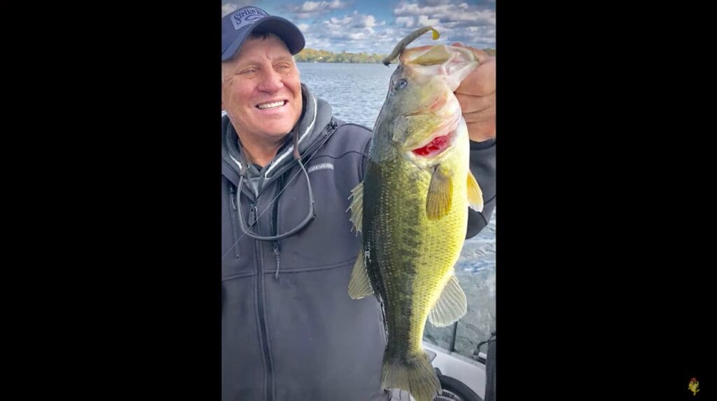 A smiling person wearing a cap and jacket holds up a large bass fish with one hand on a fishing boat, water and a cloudy sky in the background.