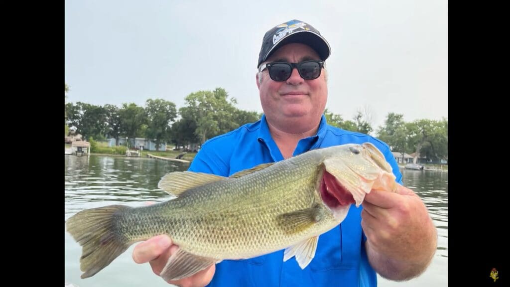 A man in a blue shirt and sunglasses smiles while holding a large summer bass with both hands, standing on a boat with water and trees in the background.