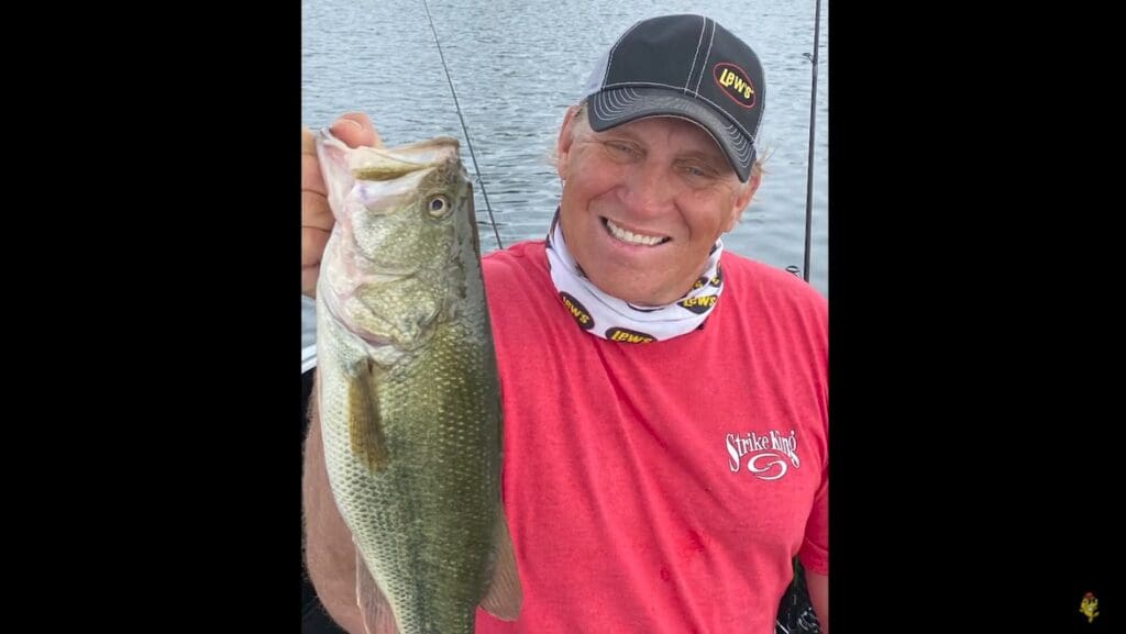 A smiling man in a red shirt and black cap holds up a large fish he caught while fishing, standing on a boat with water in the background.