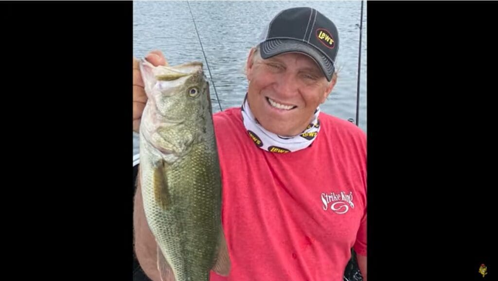 Mike, a smiling man in a red shirt and black cap, holds up a large fish in front of a lake, with fishing gear visible in the background.