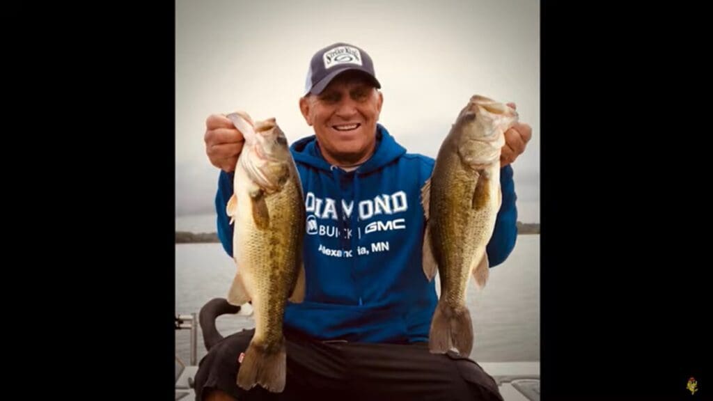 A smiling man in a blue hoodie and cap sits on a boat, holding up two large fish, one in each hand, ready to share his favorite fishing tips by the lake under a cloudy sky.