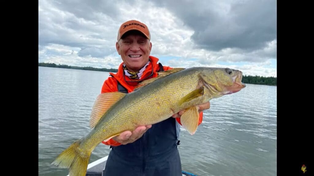 A smiling person in an orange cap and jacket holds a large yellow fish while standing on a boat, enjoying fall fishing on a lake with cloudy skies and distant trees in the background.