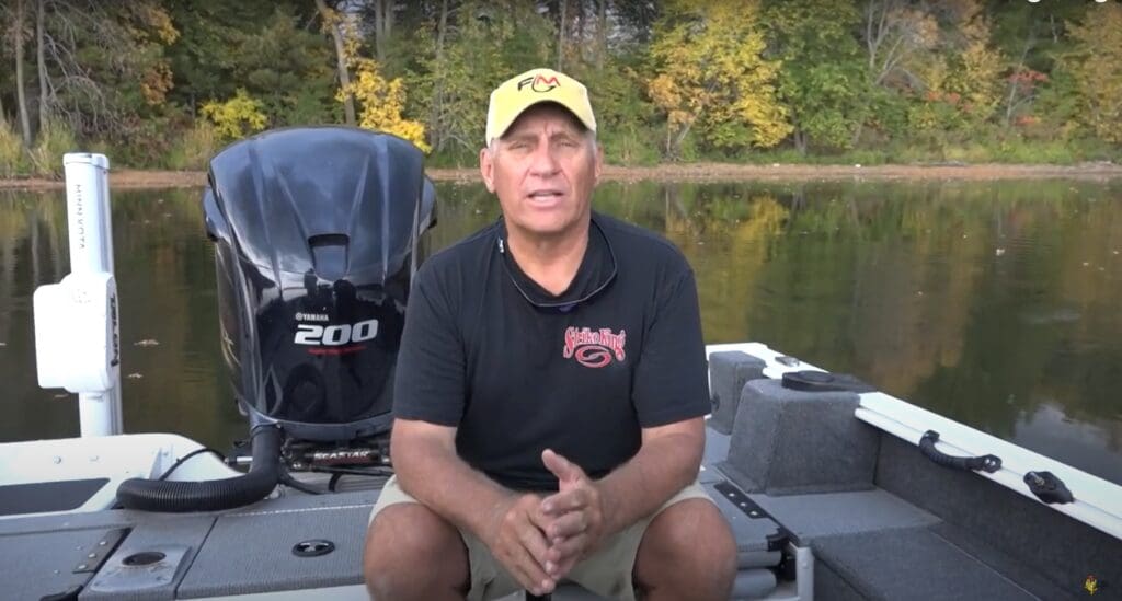 A man wearing a yellow hat and black shirt sits on a fishing boat in front of a lake, surrounded by autumn-colored trees. He faces the camera and appears to be speaking about his fishing experience.