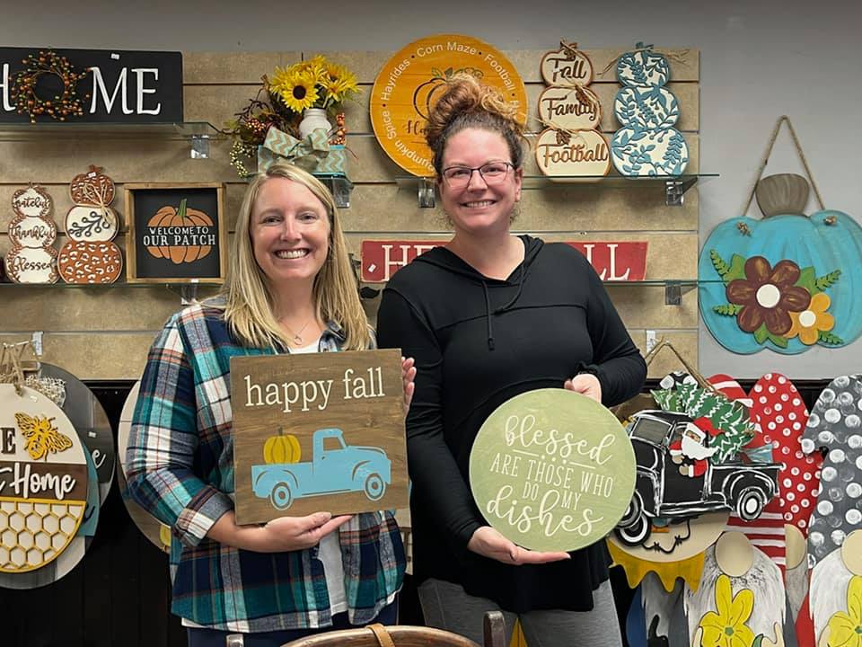 Two women smiling and holding autumn-themed wooden signs at Design Station; one sign says "happy fall" with a blue truck and pumpkin, the other says "blessed are those who do my dishes." Colorful fall decor hangs on the wall behind them.