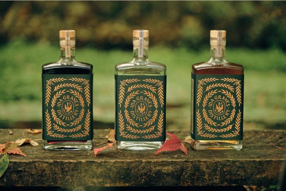 Three bottles of spirits with green and gold labels reading "MS GRANS DISTILLERS" from Ida Graves Distillery are displayed on a rustic wooden surface outdoors, autumn leaves scattered nearby and a blurred green background.