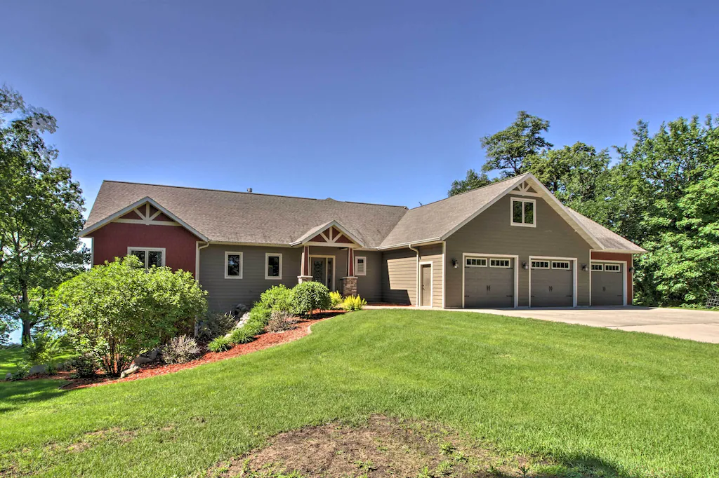 Luxury on Lake Latoka awaits in this single-story house with gray siding, a three-car garage, and a gabled roof, all nestled amid green lawns, landscaping, and trees under a clear blue sky.