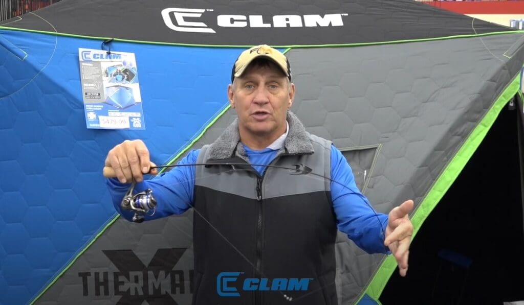 A man in a blue shirt and cap holds a fishing rod in front of a gray and blue CLAM ice fishing shelter with green trim. A product sign and the CLAM logo are visible on the shelter behind his fishing setup.