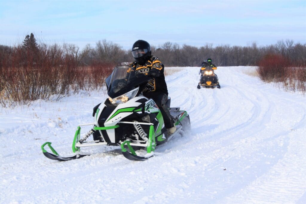 Two people ride along scenic snowmobile trails through a snowy winter landscape, with trees and shrubs in the background. The closest rider is on a green and white snowmobile, followed by another just behind.