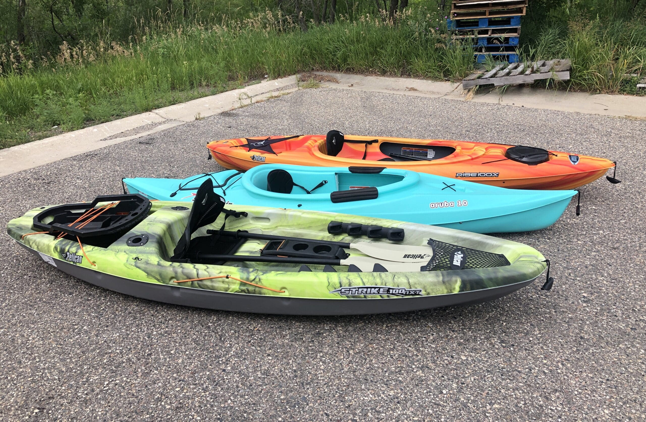 Three kayaks—one green camo, one blue, and one orange—from an Outdoor Gear Rental are parked on a gravel surface near a grassy area and wooden pallets, lined up side by side.