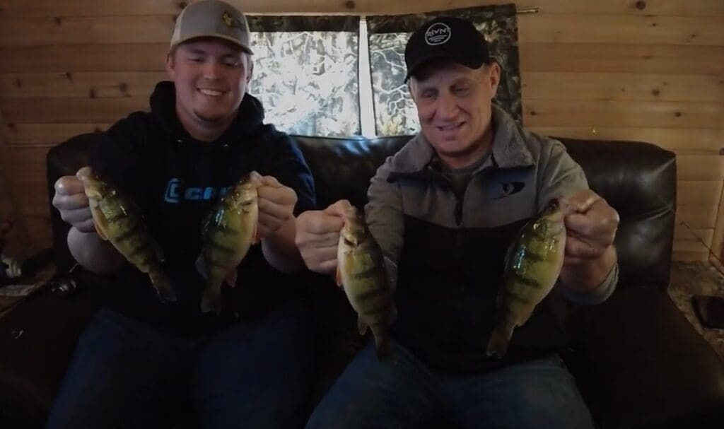 Two smiling men, including Mike Frisch, sit on a couch in a wooden cabin, each holding two fish. The man on the left wears a gray baseball cap and dark hoodie; the man on the right has a black cap and gray jacket.