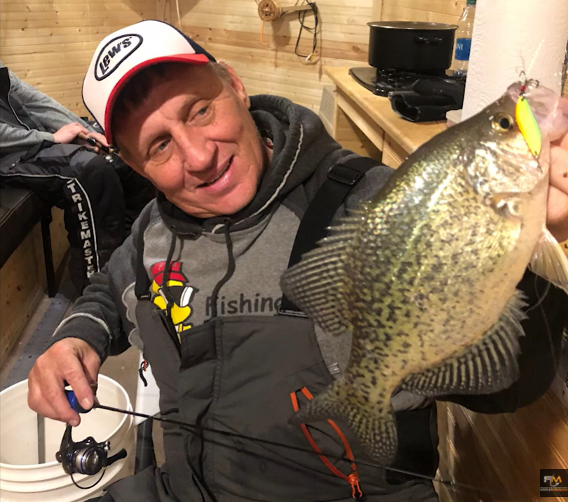 A man in a fishing outfit proudly holds up a large crappie fish with a yellow lure in its mouth, smiling inside a cozy wooden cabin. A fishing rod and bucket highlight the joy of his successful fishing adventure.