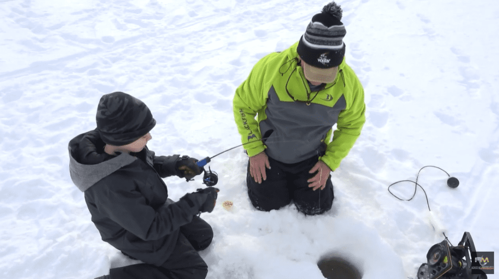 Two people dressed in winter clothing kneel on snow next to an ice-fishing hole. One person holds a small fishing rod while the other watches, enjoying a peaceful fishing moment. Ice fishing equipment is nearby.