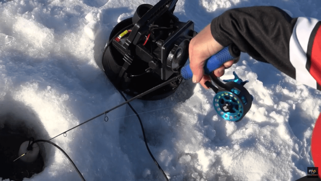 A person holds a blue-handled fishing rod over a hole in the ice, next to a black reel and other ice fishing equipment scattered on the snowy surface.