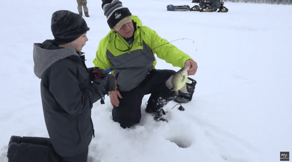 A man kneels on snow by an ice fishing hole, proudly holding up his catch, while a child in winter clothing stands beside him. Fish, snowmobiles, and gear dot the snowy landscape in the background.
