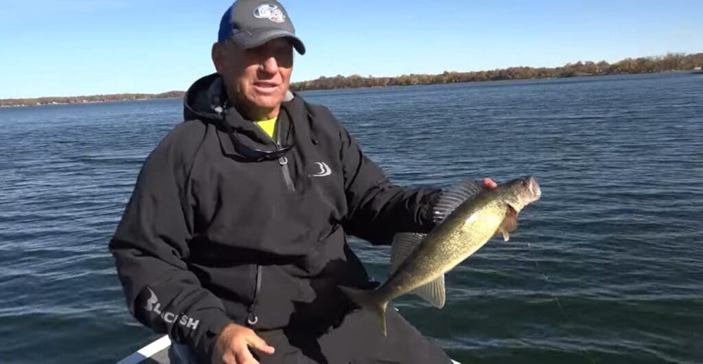A man in a black jacket and baseball cap sits on a boat during the fishing opener, holding a large fish he caught, with a calm lake and distant tree-lined shore in the background under a clear blue sky.
