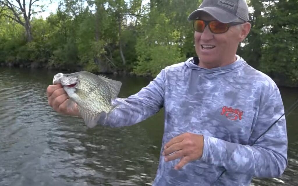 A man wearing sunglasses and a blue camo hoodie holds a large crappie by the water, smiling, with trees in the background—showing off one of the finest crappies of the day.