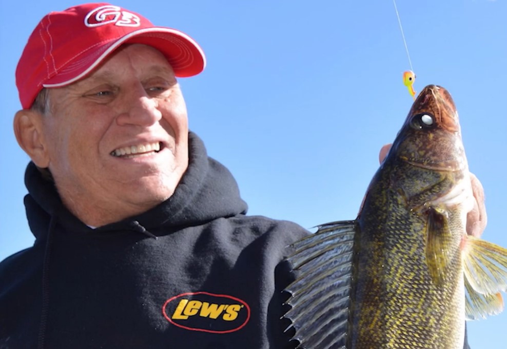 A smiling man in a red cap and black Lew's hoodie holds up a large walleye on a fishing line against a clear blue sky.