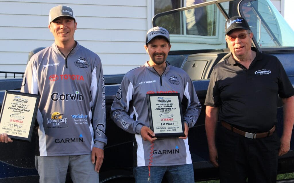 Three men stand in front of a boat. Two on the left wear fishing shirts and hats, smiling and holding 1st place walleye plaques. The man on the right wears sunglasses, a cap, and a black polo shirt, also smiling.