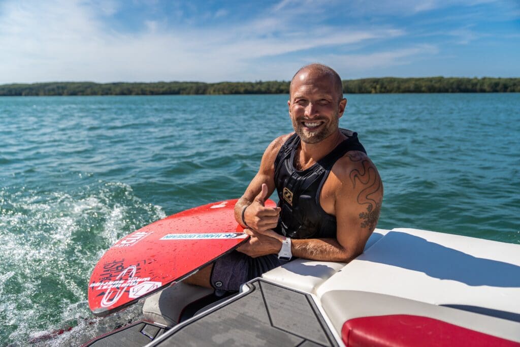 A smiling man in a black life vest sits on the edge of a boat holding a red wakeboard, ready for water sports, with blue water and a distant treeline in the background on a sunny day.