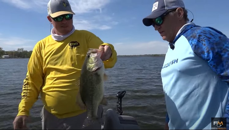 Two men stand on a boat in the water; one, wearing a yellow shirt and hat, proudly holds up a large mouth bass while the other, in a blue shirt and cap, admires the impressive catch. Trees and shoreline are visible in the background.