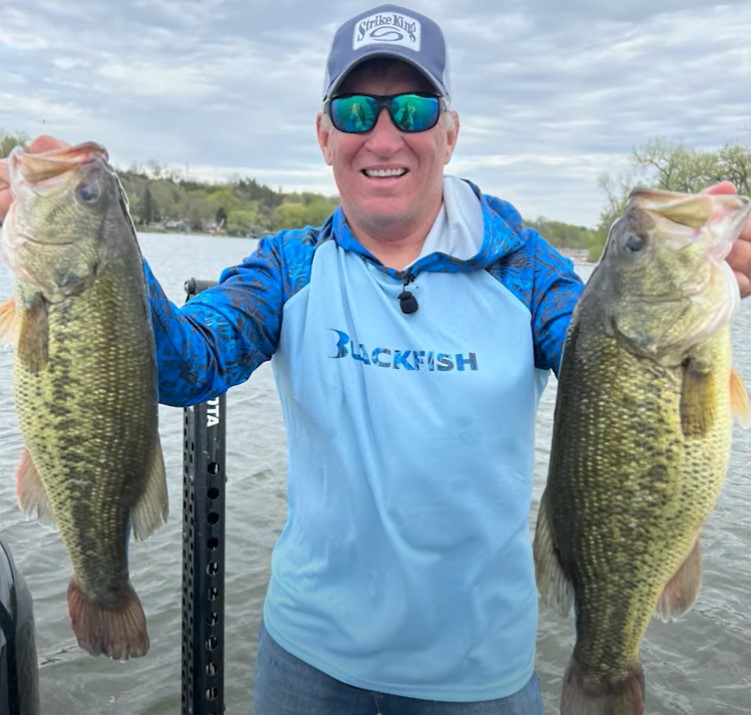 A smiling man in sunglasses and a blue shirt stands on a boat, holding up two large bass fish caught with a squarebill, with water and trees in the background.