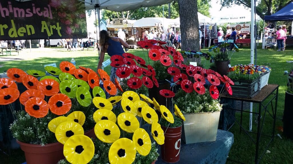 Colorful ceramic flowers in red, orange, and yellow fill pails at an outdoor Art in the Park fair. A sign reads “Whimsical Garden Art” as people wander among tents and booths in the lively background.