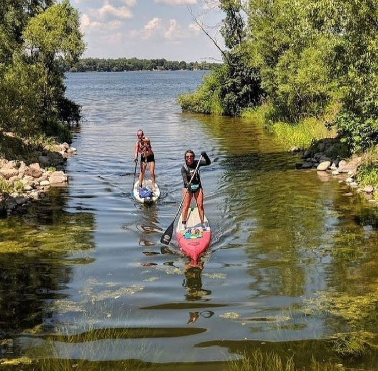 Two people paddleboarding on a calm, narrow waterway surrounded by green trees and grass, heading toward an open lake under a blue sky with scattered clouds during the Big Ole Paddlefest event.