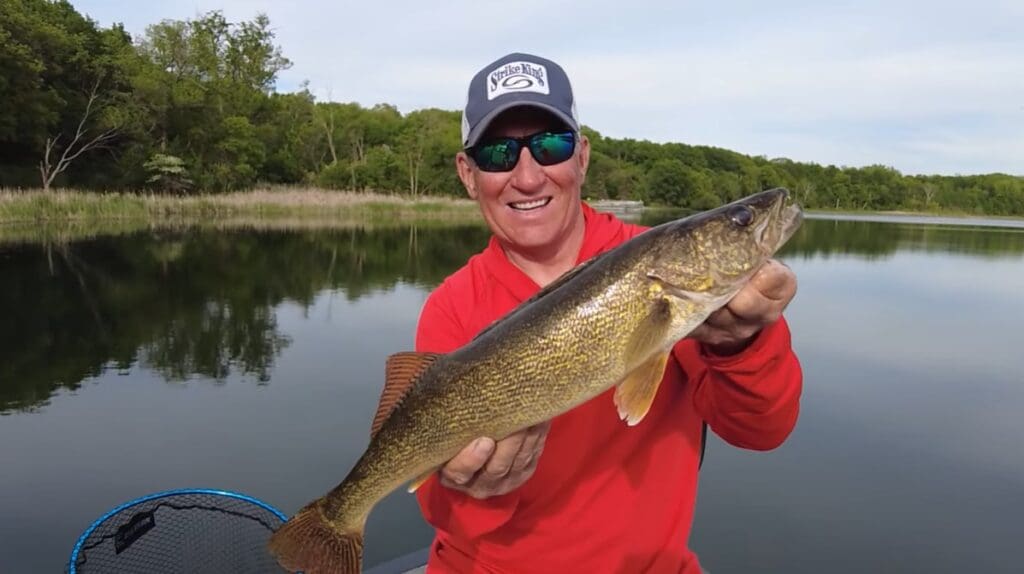 A man in a red shirt and cap smiles while holding a big fish, standing proudly in a boat on a calm lake with trees in the background.