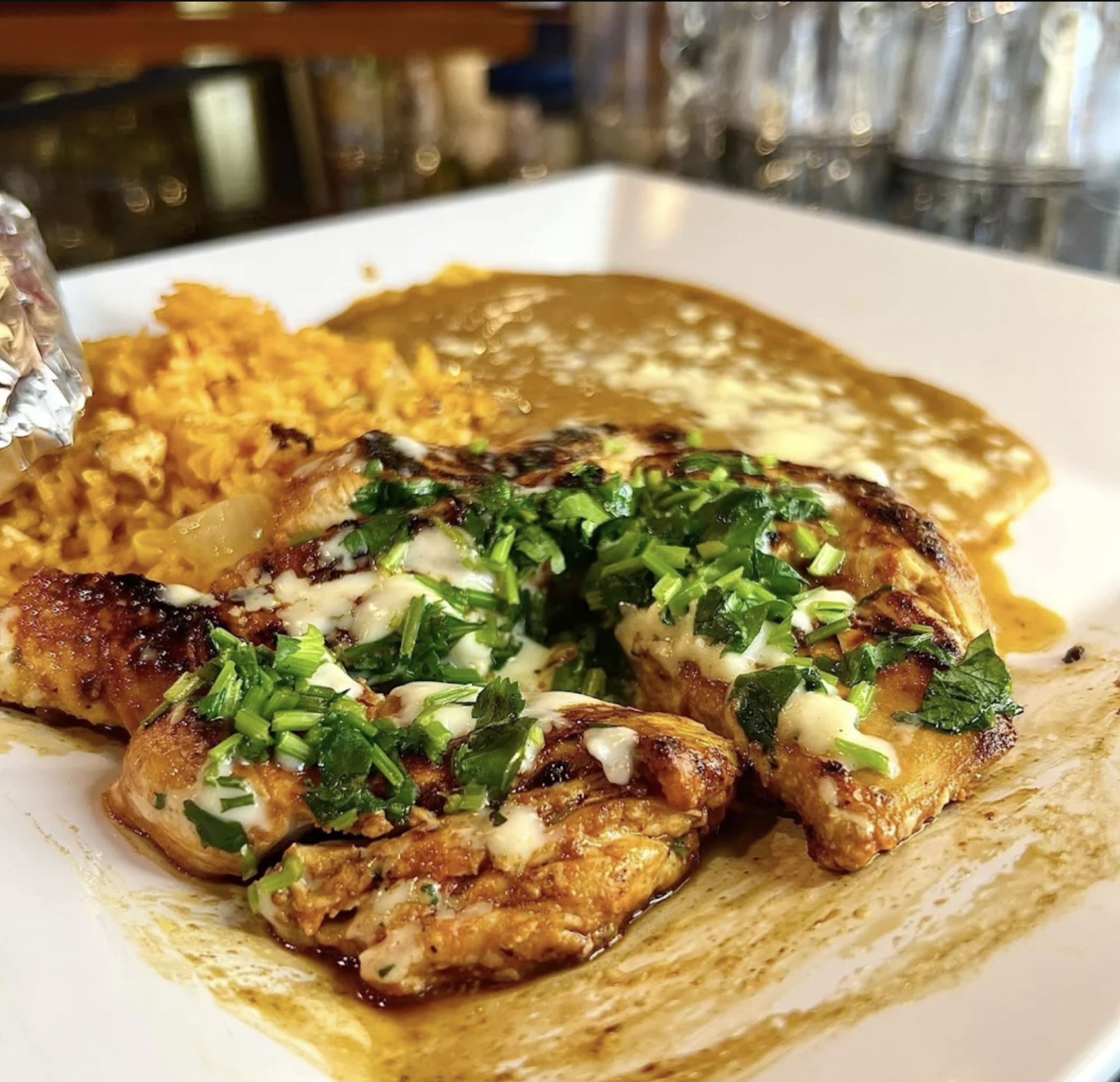 Grilled chicken breast topped with chopped herbs and melted cheese, served with yellow rice and a creamy brown sauce on a white plate—a flavorful El Loro specialty. Glassware is visible in the background.