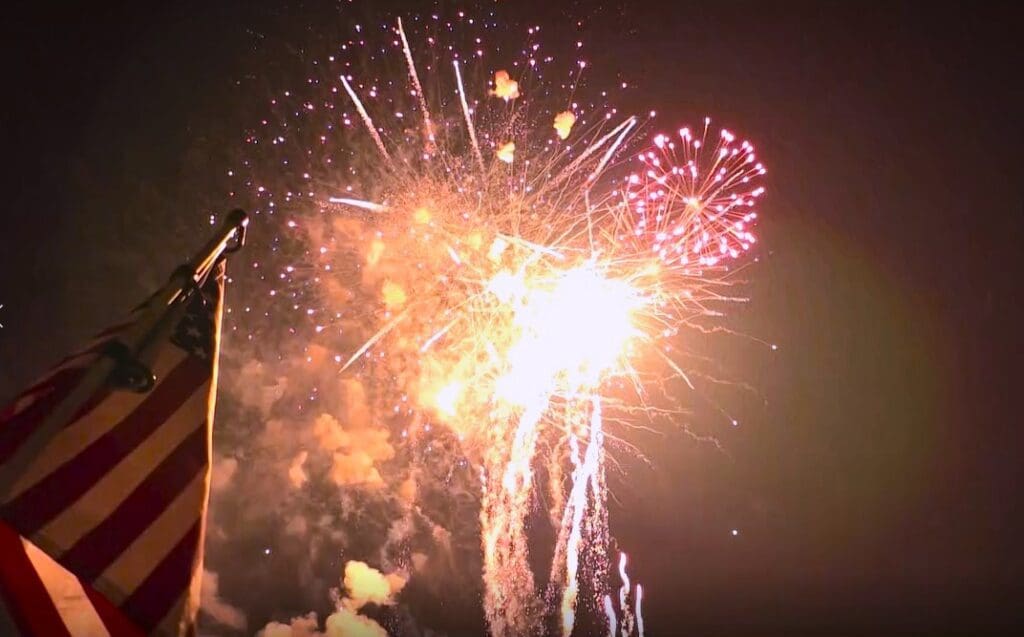 A brightly lit night sky filled with colorful fireworks. In the foreground, part of an American flag is visible, suggesting a festive or patriotic 4th of July celebration in the lakes area.