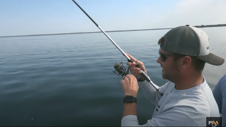 A man wearing a gray cap and sunglasses is holding a fishing rod over calm water, appearing to reel in a catch with swimbaits on a sunny day.