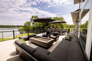 Spacious outdoor patio with modern black furniture, a fire pit, umbrellas, and a dining area—this Alexandria Dream Home overlooks a lake and is surrounded by lush greenery under a partly cloudy sky.