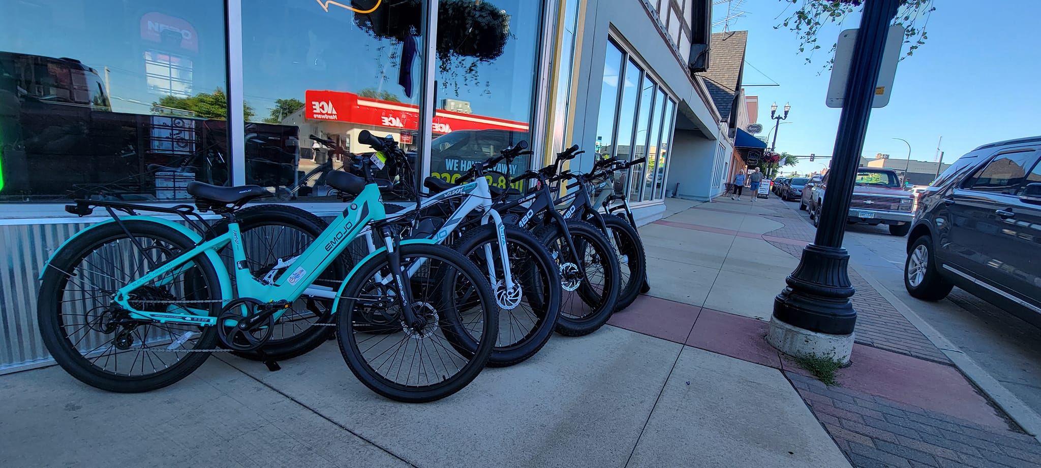 Several bicycles, including some from Central Ebikes, are lined up and parked on a sidewalk outside a storefront, with cars parked along the street and buildings visible in the background.