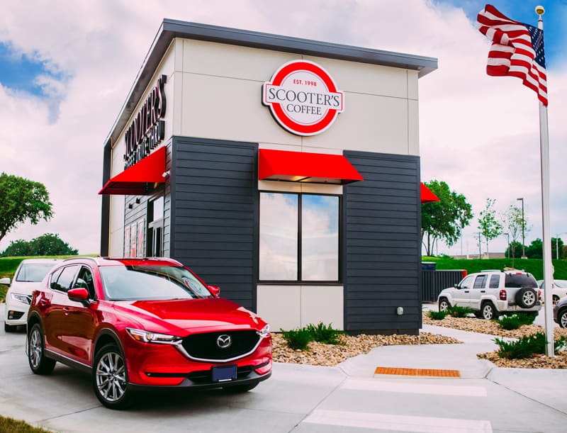 A red SUV waits at the drive-thru of Scooter’s Coffee, a modern café with bold red accents. The American flag waves nearby, while greenery surrounds Scooter's Coffee under a partly cloudy sky.