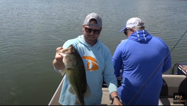 Two people on a boat; one man in sunglasses and a light blue hoodie, proudly holding up a big fish and smiling at the camera, while the other in a blue shirt faces away, fishing on a calm lake.