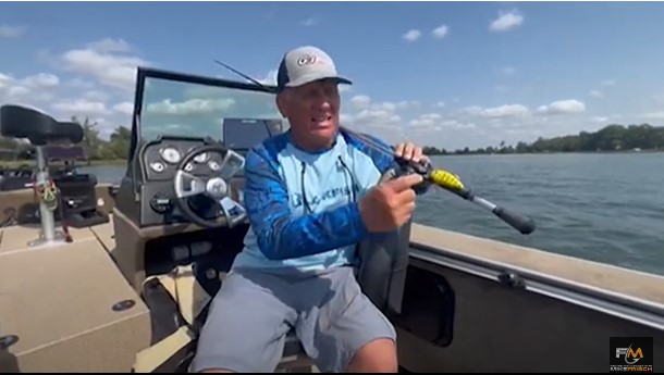 A man in a blue long-sleeve shirt and cap sits in a boat on a lake, holding a fishing rod with moving baits. The sky is clear with scattered clouds, and trees are visible in the background.