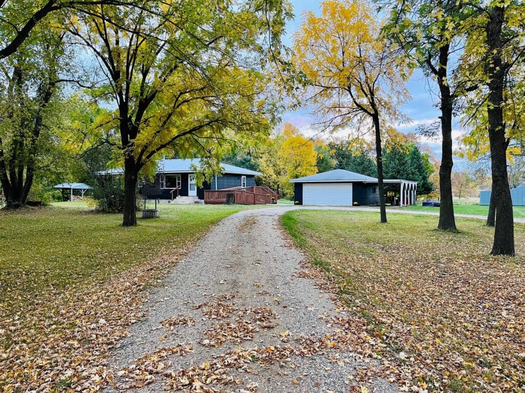 A gravel driveway leads to a one-story Countryside Oasis with a porch and detached garage, surrounded by trees in autumn foliage and a lawn covered in fallen leaves.