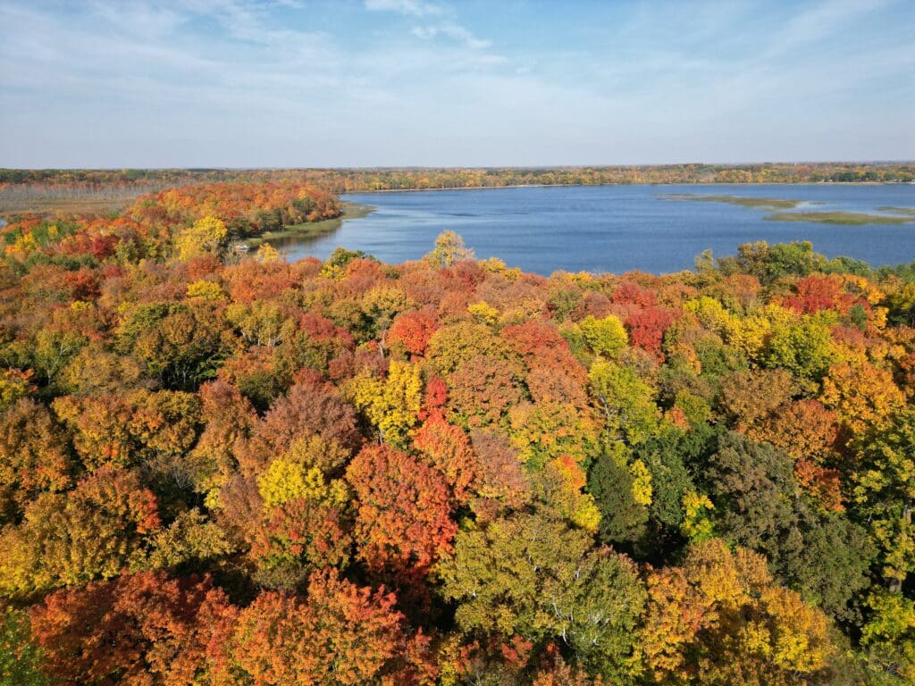Aerial view of a forest with vibrant autumn foliage in shades of red, orange, and yellow surrounds the serene blue lake near McCabe Cabin, all beneath a partly cloudy sky.