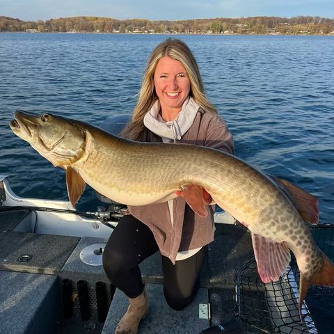 A smiling woman on a boat holds a large musky over her knees. The boat floats on a blue lake with a wooded shoreline in the background beneath a partly cloudy sky.