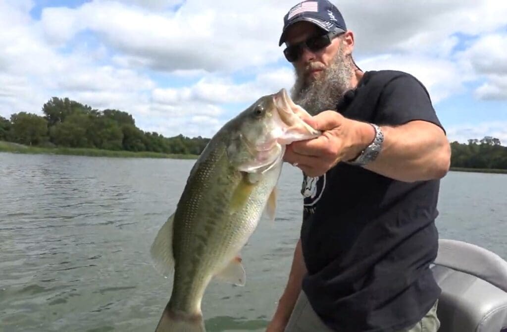 A bearded man wearing sunglasses and a cap holds a large fish on a boat, enjoying some fall fishing, with a lake and trees in the background under a partly cloudy sky.