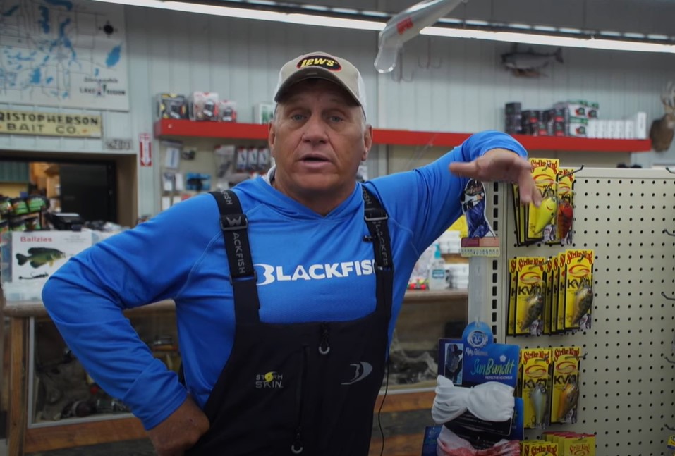A man in a blue "Blackfish" shirt and overalls leans on a store display stocked with bass fishing lures. He wears a white cap and stands in a fishing supply shop filled with gear, signs, and mounted fish on the walls.