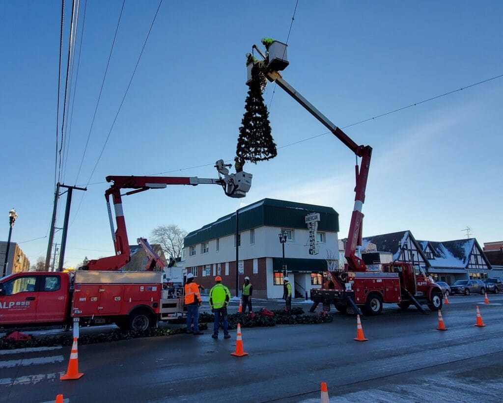 Workers in safety gear use bucket trucks to hang a large Christmas tree decoration above the street for the Lights on Broadway event, with orange cones marking the work area. A two-story building and houses are visible in the background.