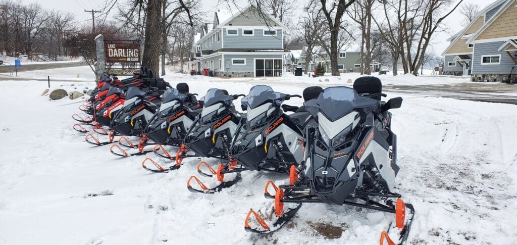 A row of snowmobiles with orange skis, available for rentals at Lake Darling Resort, is parked on snow beside a sign that says "Lake Darling Resort." Snow-covered trees and houses are visible in the background.