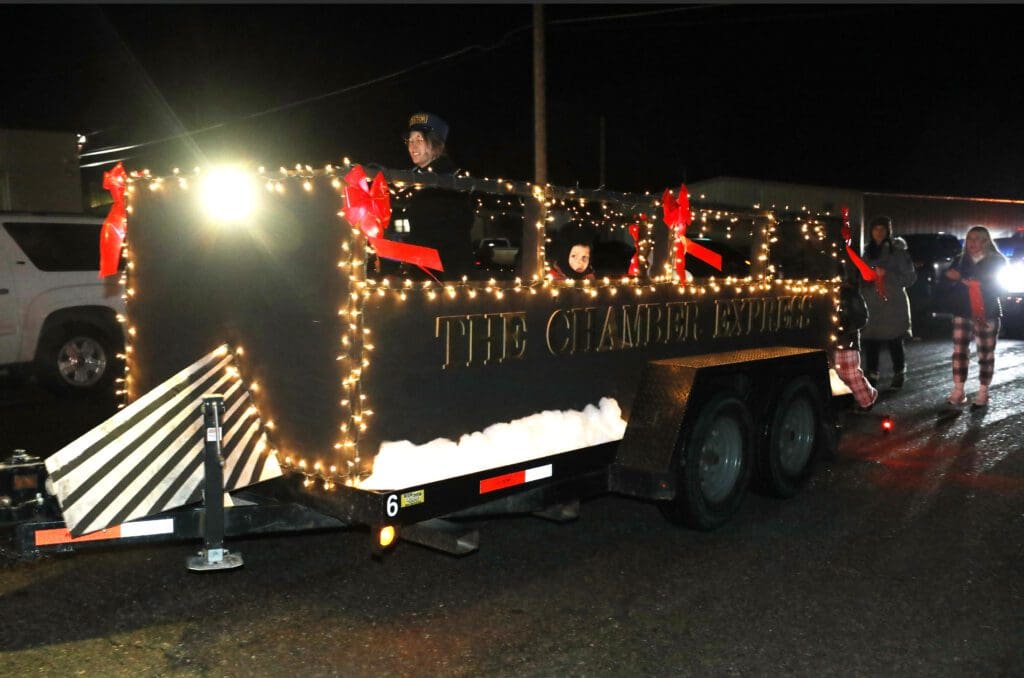 A parade float decorated as a black train car with "THE CHAMBER EXPRESS" in gold letters, festive lights, red bows, and people inside celebrates Christmas in the Fort as it moves down the street at night.