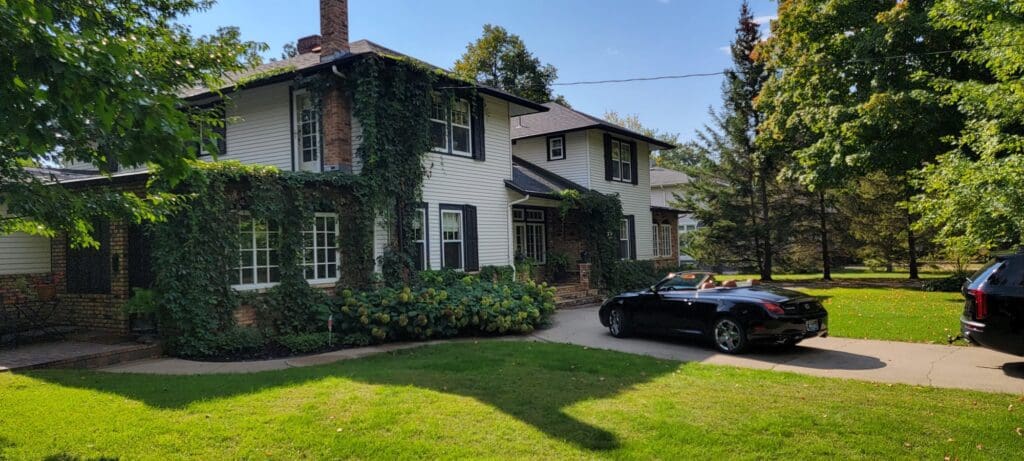 A two-story house with white siding and brick accents, partially covered in ivy, stands near the Jones Big Top. A black convertible is parked in the driveway, while the yard is shaded by green trees under a clear blue sky.